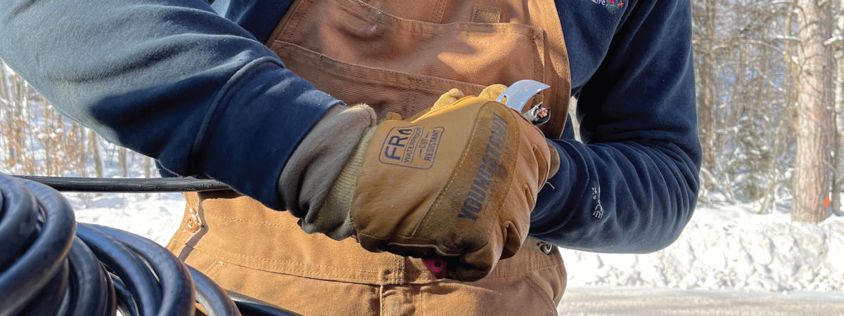 A close-up of a person outdoors wearing a brown work apron over a navy blue long-sleeve shirt, with tan work gloves on. Snow and bare trees are visible in the blurred background.