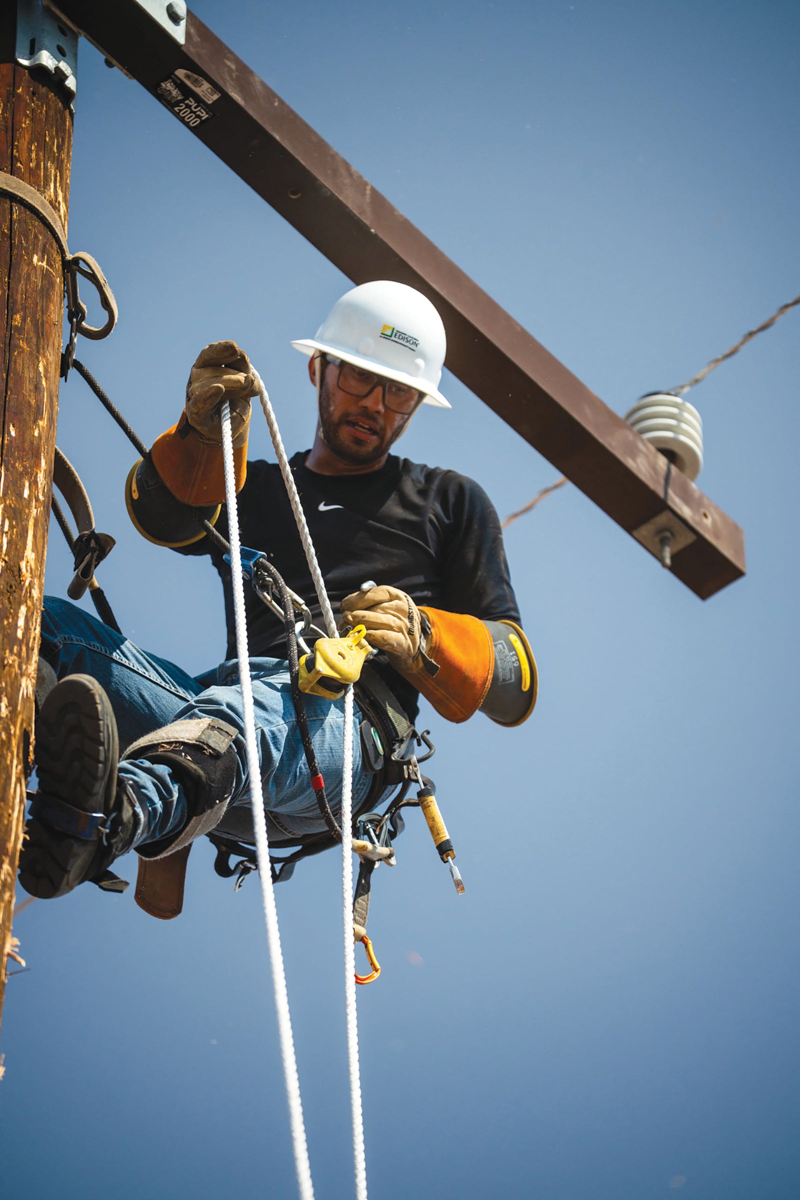 A utility worker suspended in a harness on a utility pole, wearing a white hard hat, black shirt, blue jeans, and yellow Youngstown gloves against a clear blue sky.