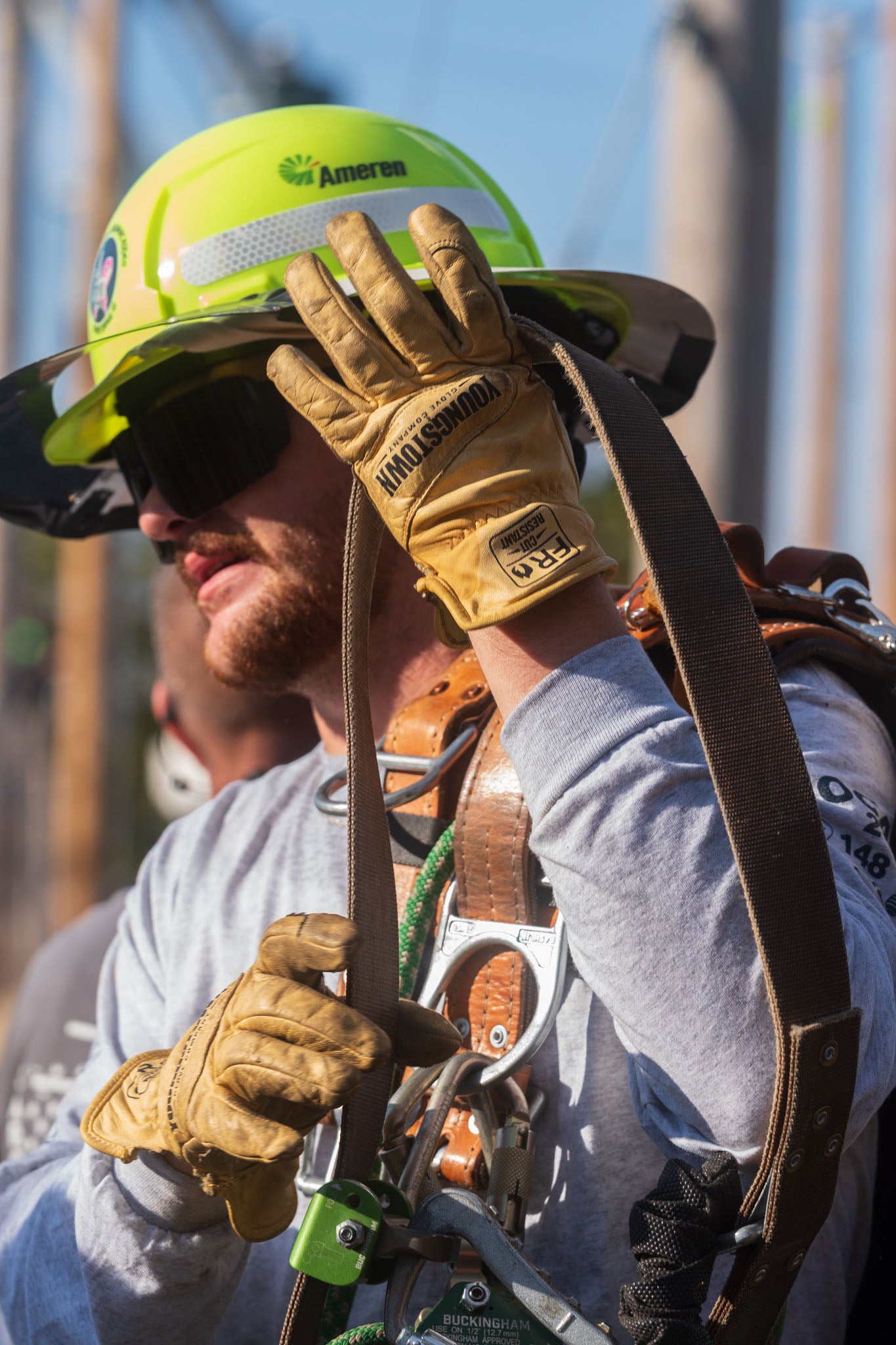 A close-up lifestyle shot of a lineman wearing a bright green hard hat, Youngstown leather work gloves, and a climbing harness with a brown strap, adjusting his helmet outdoors.
