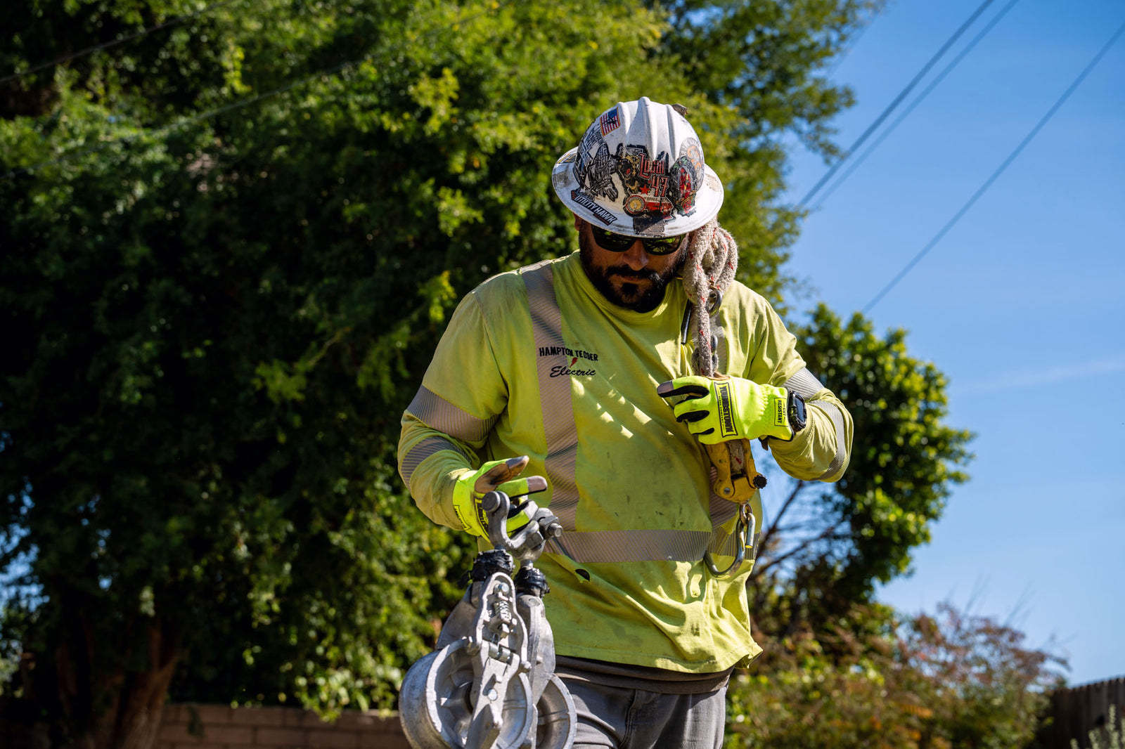 A man wearing a white hard hat, a high-visibility yellow work shirt, and Youngstown gloves holds a metal climbing or safety accessory outdoors against a background of green trees and blue sky.
