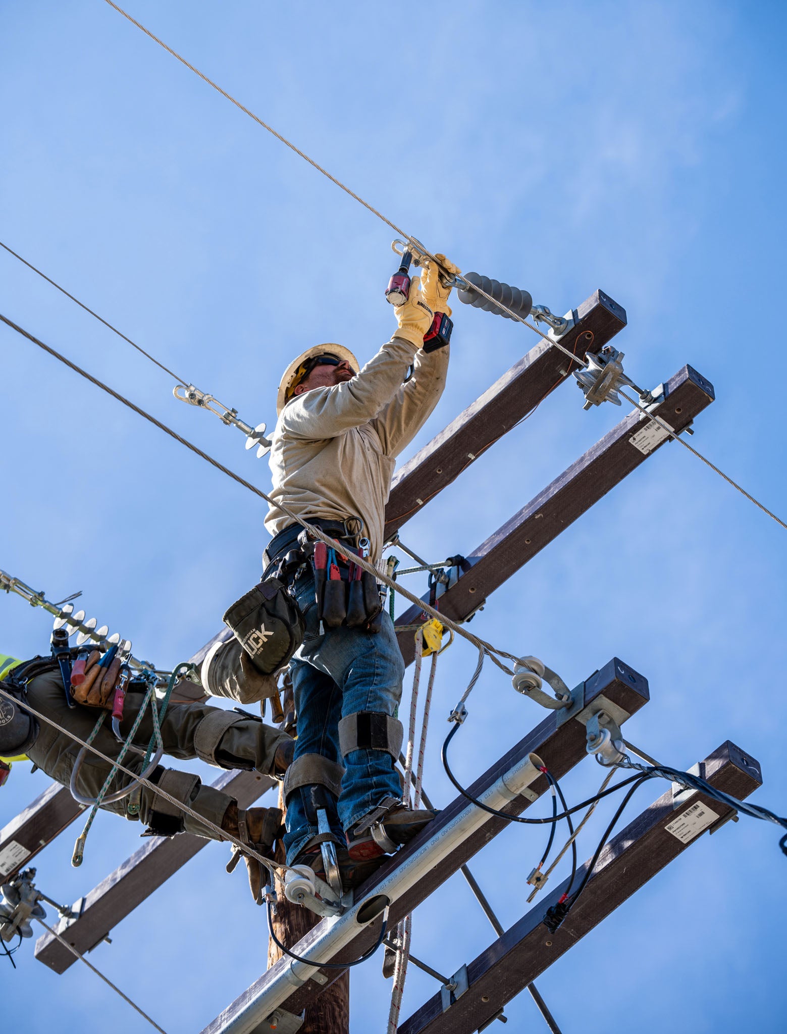 Utility workers wearing Youngstown gloves, safety gear, and harnesses climb a wooden utility pole against a clear blue sky, handling overhead power lines.