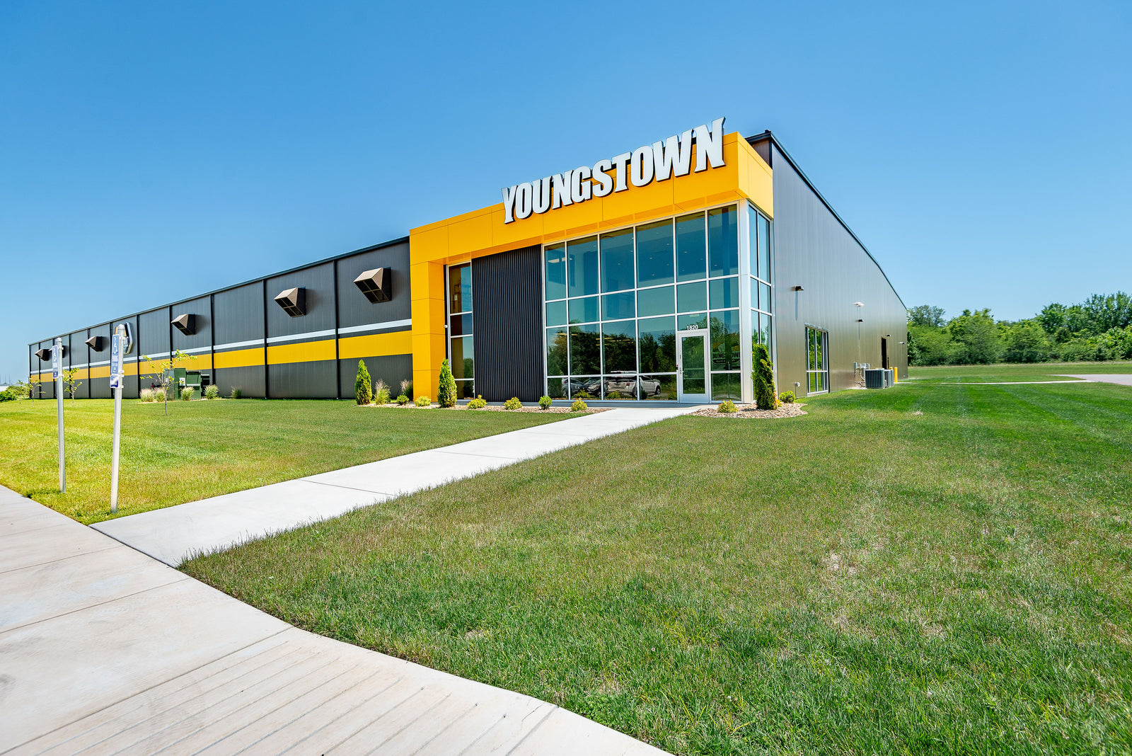 A modern warehouse building with a bold yellow and dark gray facade, featuring large glass windows and a prominent 'YOUNGSTOWN' sign on the exterior, set against a clear blue sky and green grass.